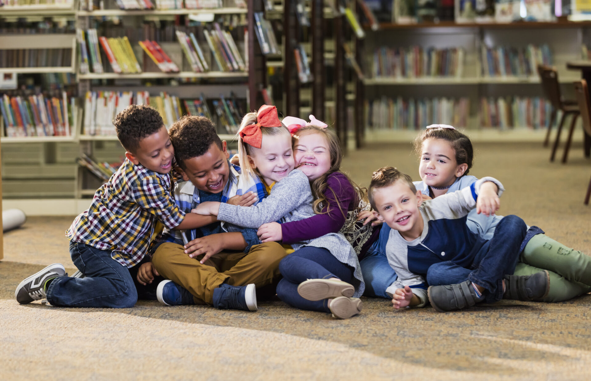 A group of six multi-ethnic children sitting together on the floor of a school library, hugging. The girl sitting 3rd from the right, has down syndrome.