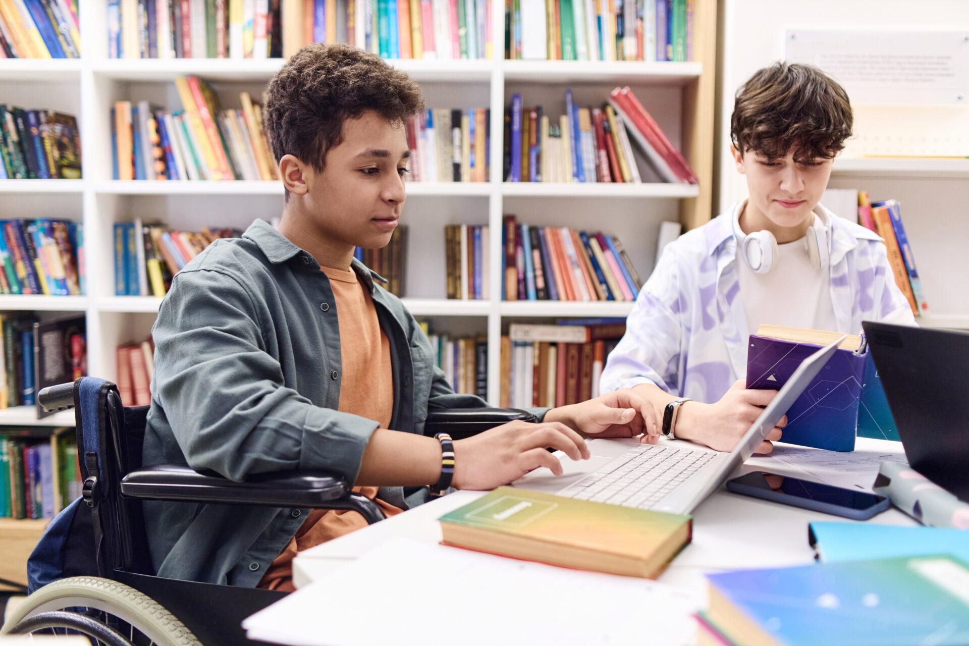 Side view portrait of young teenage boy with disability using laptop computer in school library with group of students.