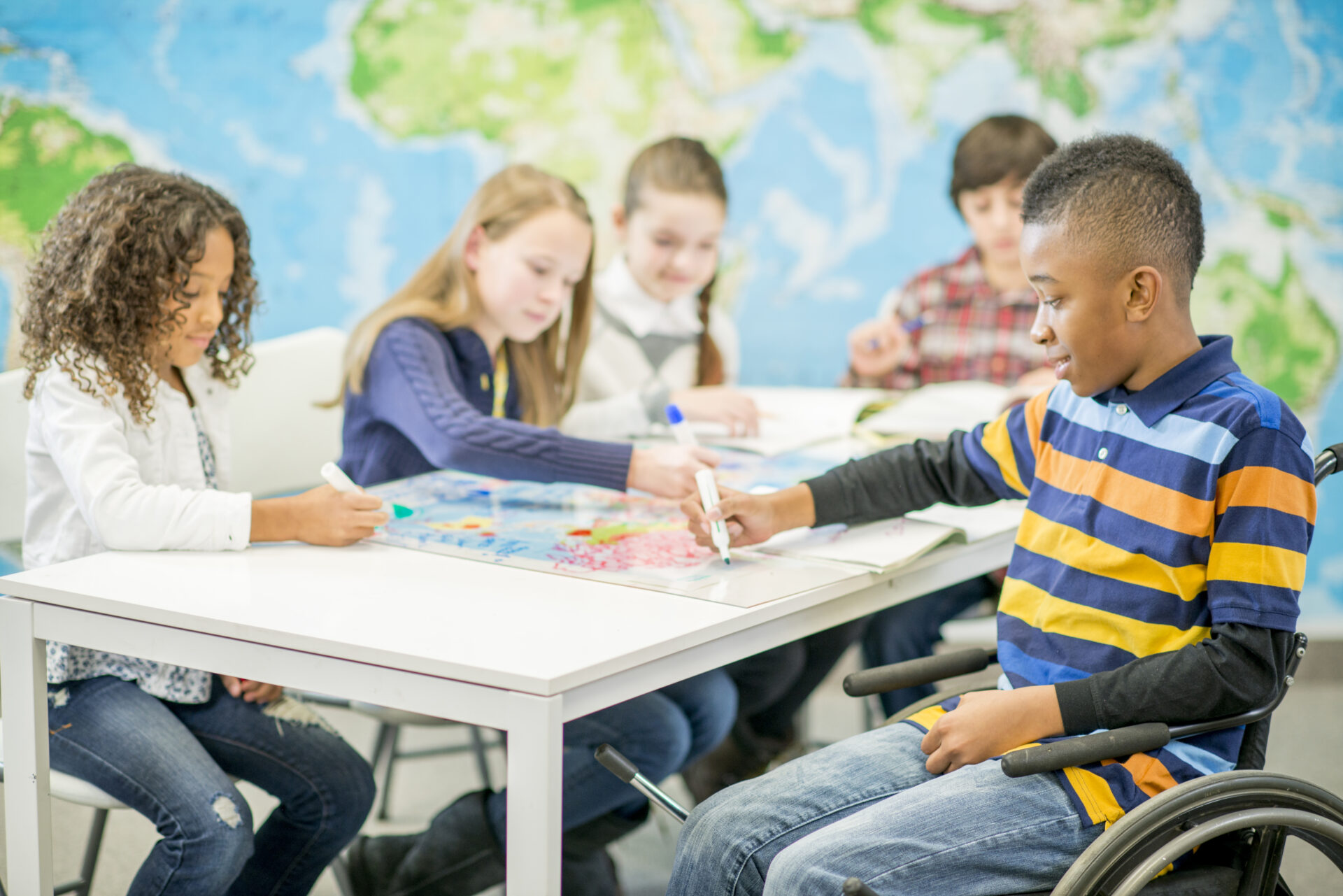 A multi-ethnic group of elementary age children are sitting at a desk and are drawing on a world map for geography class. One handicap child is sitting in a wheel chair.